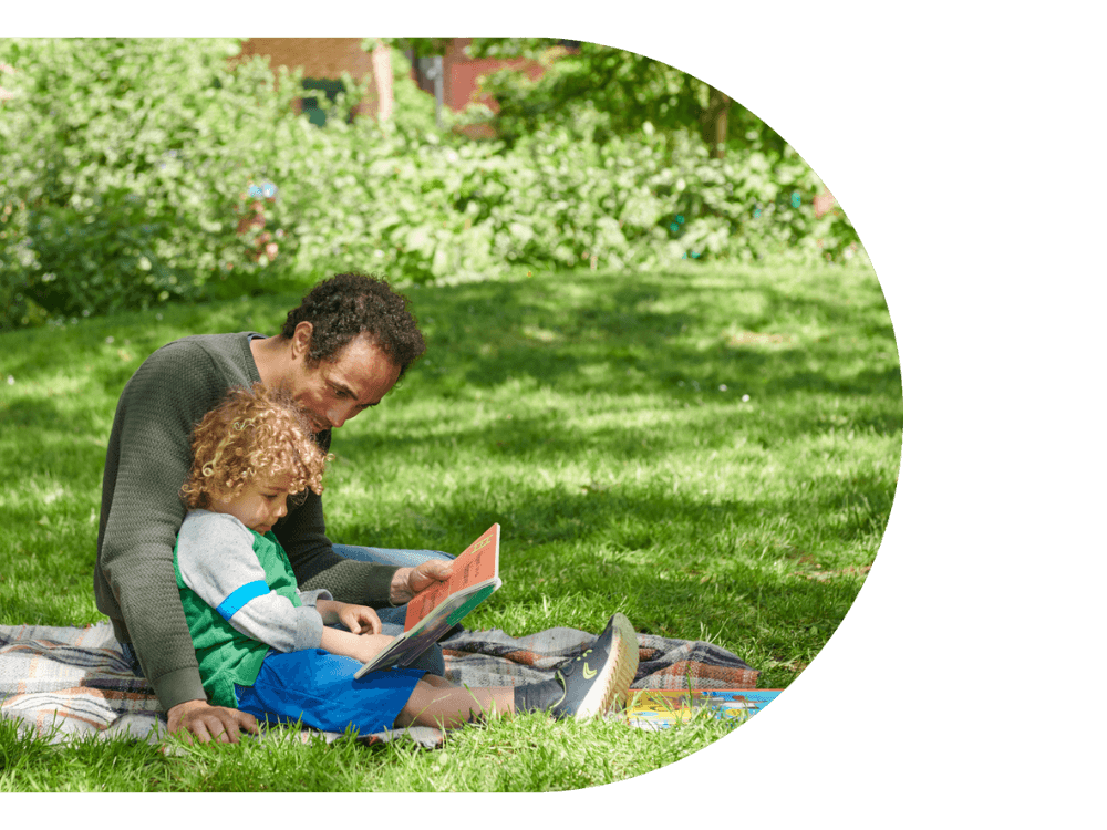 A man reading a book to his child sitting in the grass at QEOP.