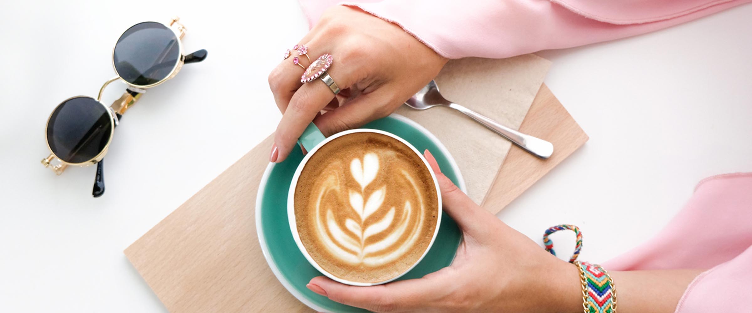Hands around a mug of coffee with sunglasses on the table next to the mug.